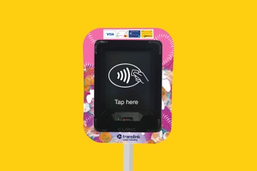 An onboard validator. The screen is showing the words 'Tap here' and the contactless payment symbol. The screen is surrounded by a bright pink decal, with card payment options illustrated at the top (Visa, Mastercard, Amex and go card).